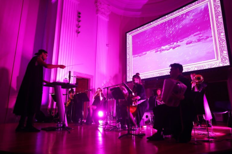 Concert en directe amb orquestra davant d’una pantalla, sota llums de color magenta