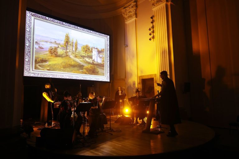 Orquestra tocant en directe a l’escenari, sota una pantalla que projecta una pintura de paisatge